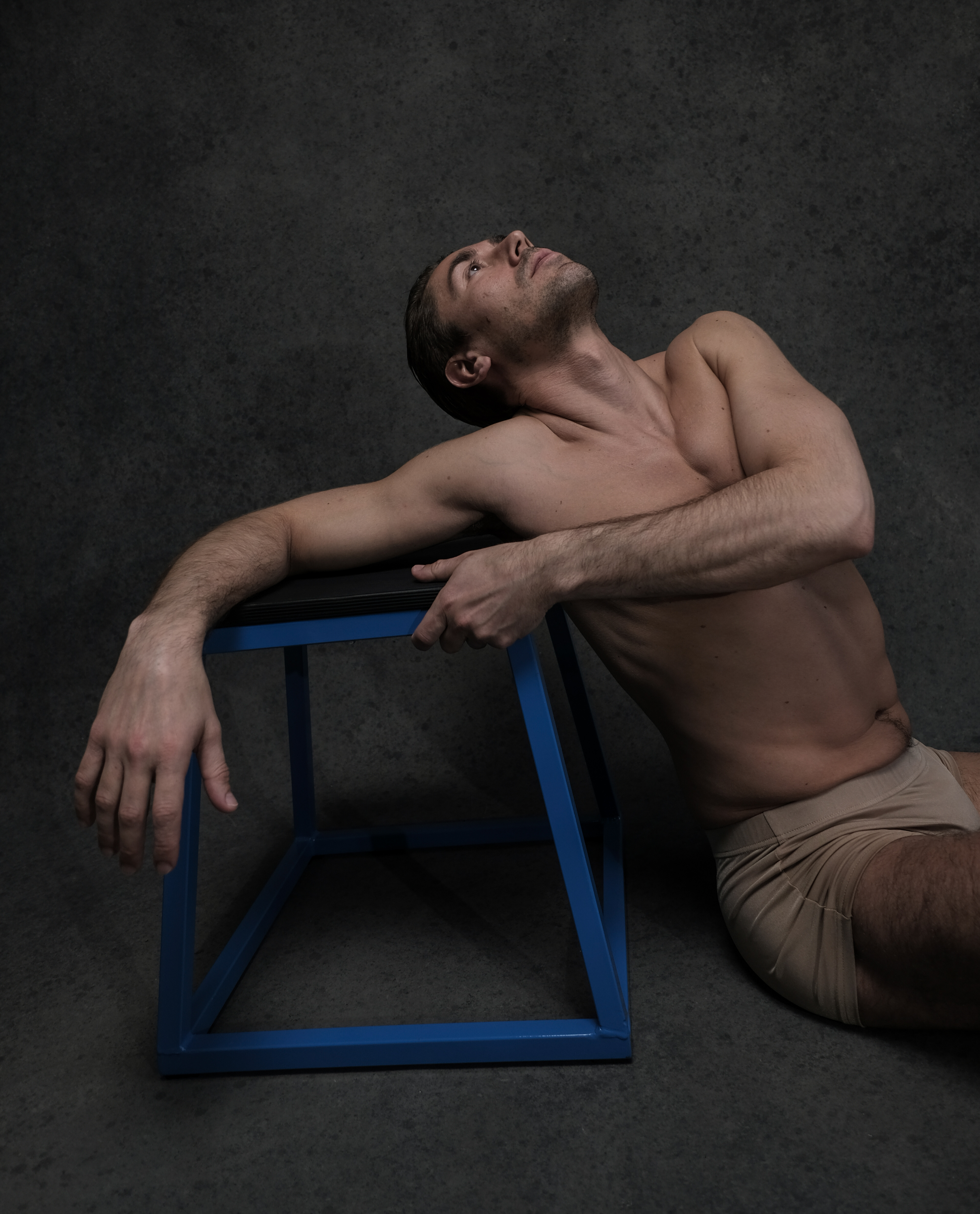a shirtless male dancer leans on a blue metal stool in a relaxed pose, his head tilted back and eyes closed. he is positioned against a dark gray textured background. the lighting is soft and natural, highlighting the contours of his body.