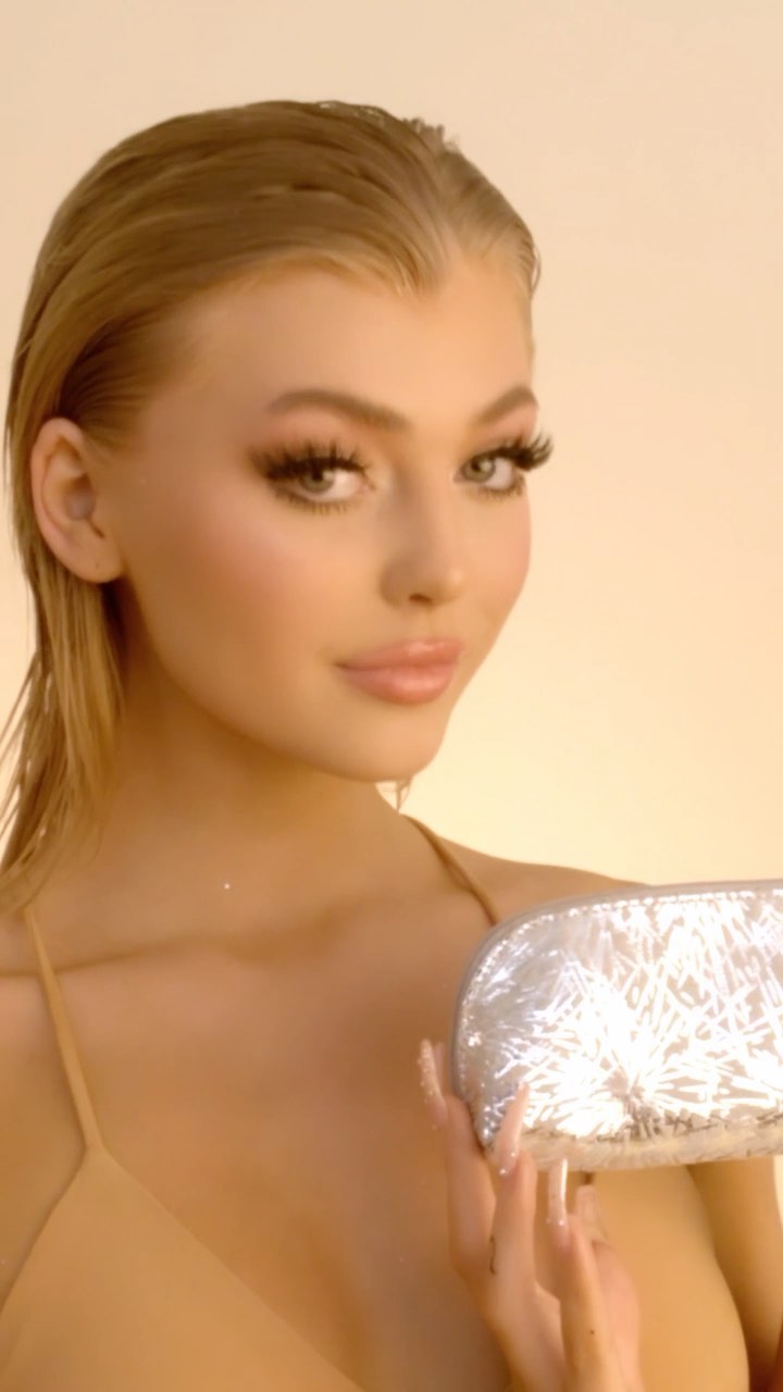 a closeup shot features a woman with blonde hair styled in a sleek, wet look. she has a full face of makeup, including long eyelashes, and is holding a silver makeup bag with intricate designs in her hand. the background is a soft, creamy color, which complements her tan skin tone. she's wearing a simple strap top in a nude shade, allowing the focus to remain on her face and the makeup bag. she poses confidently while looking into the camera.