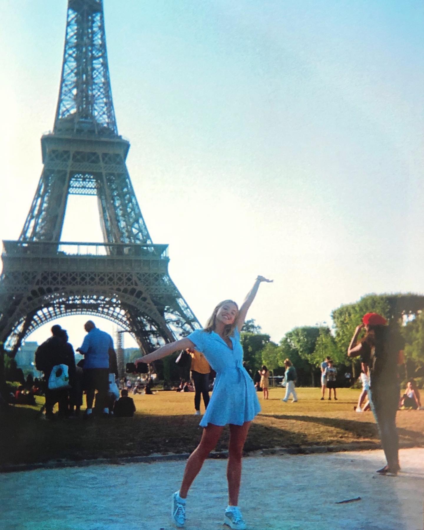 a woman in a blue dress poses in front of the eiffel tower in paris, with her arms outstretched. there are other people in the park around her, and the sky is clear.