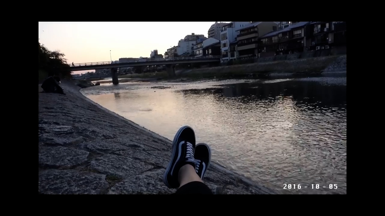 the image shows a firstperson perspective of someone sitting by a river in an urban environment. the person's feet are propped up, wearing black and white vans shoes. the river flows from left to right, reflecting the soft, evening light of the setting sun. a bridge spans the river in the background, connecting buildings on either side. the overall atmosphere is one of relaxation and tranquility.  the image appears to be taken in japan.
