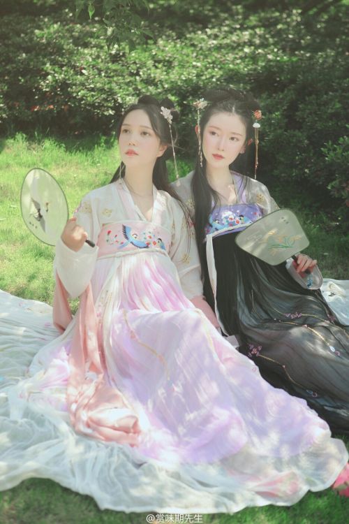 two women dressed in hanfu are sitting outside on a blanket. the woman on the left is wearing a white and pink hanfu and holding a fan in her right hand. the woman on the right is wearing a grey and black hanfu and holding a fan. they are sitting on a white blanket in a garden with greenery.