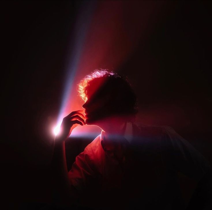 a silhouette of a man with red hair is lit from the right side, casting a shadow over his face and body. the light is warm and inviting, while the background is dark and mysterious. the man's hand rests on his chin, as if he is in deep thought or contemplation. the overall effect is moody and dramatic, with a touch of artistic beauty.