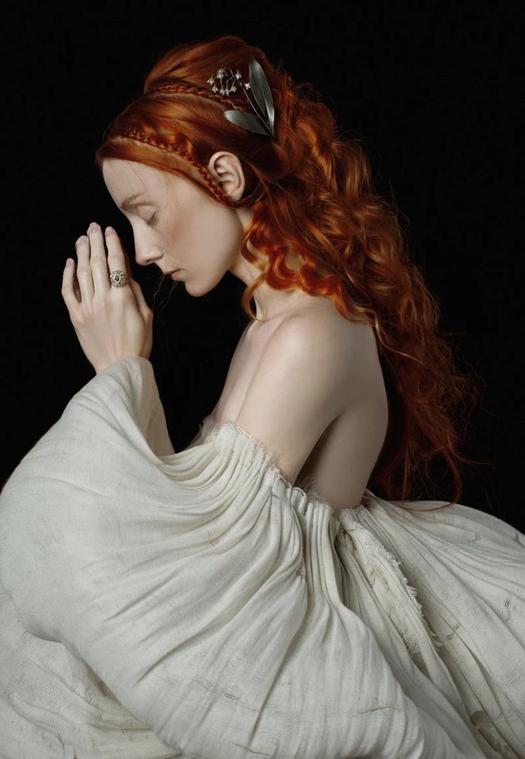 a woman with long red curly hair wears a white vintagestyle dress.  she is looking down with her hands clasped together.  the background is black.