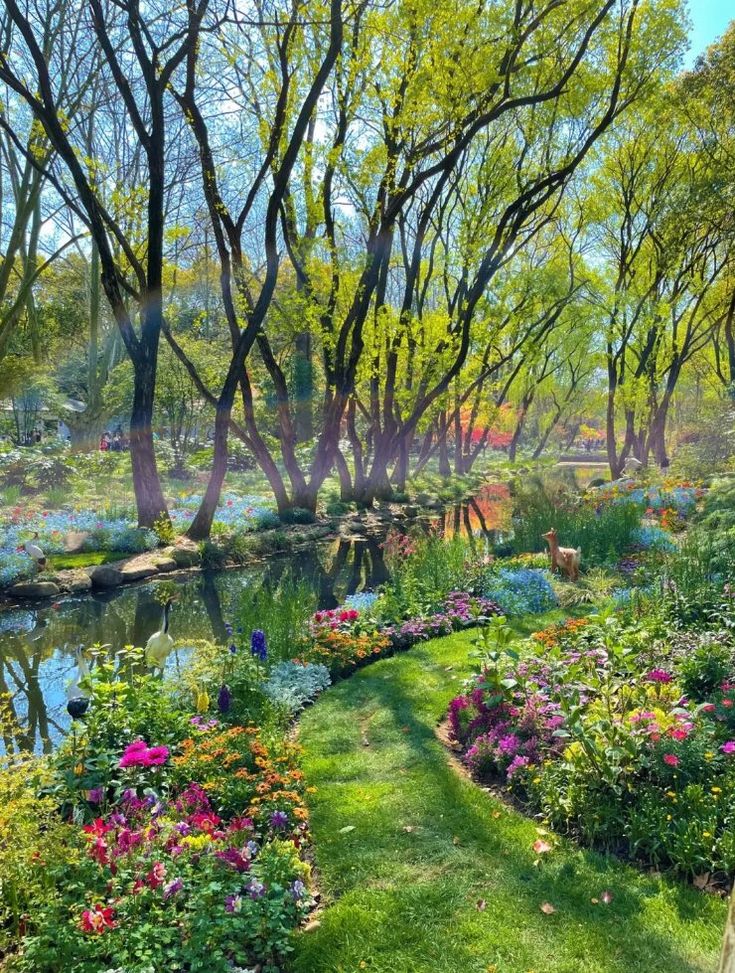 the image depicts a lush and vibrant garden scene. a narrow stream reflects the sky and surrounding trees, with a goose visible near the water's edge. a grassy path winds through colorful flowerbeds filled with various blooms. trees line the stream, their branches extending overhead, casting dappled shadows on the scene. a deer figurine stands among the flowers. the overall impression is one of tranquility and natural beauty.
