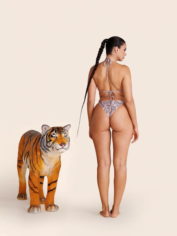 a woman with long, dark hair styled in a ponytail stands with her back to the camera, showcasing a twopiece animal print bikini. she stands against a minimalist cream background beside a lifesized tiger statue facing left. the scene is brightly lit in a professional studio setting.