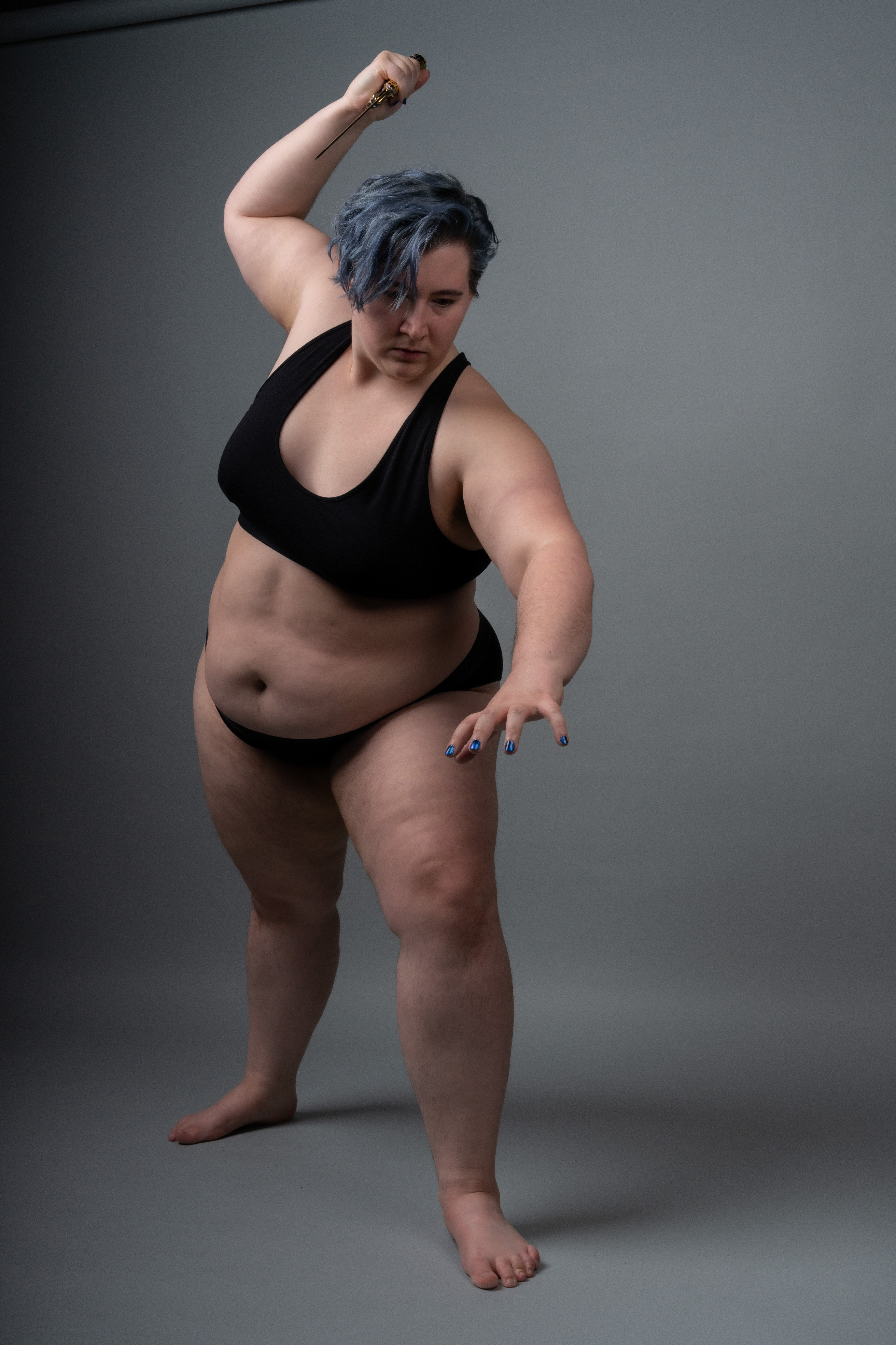 a plussize woman with blue hair is striking a combat pose with a sword in a studio setting. she is wearing a black bra and panties, and is barefoot. the background is a solid grey.