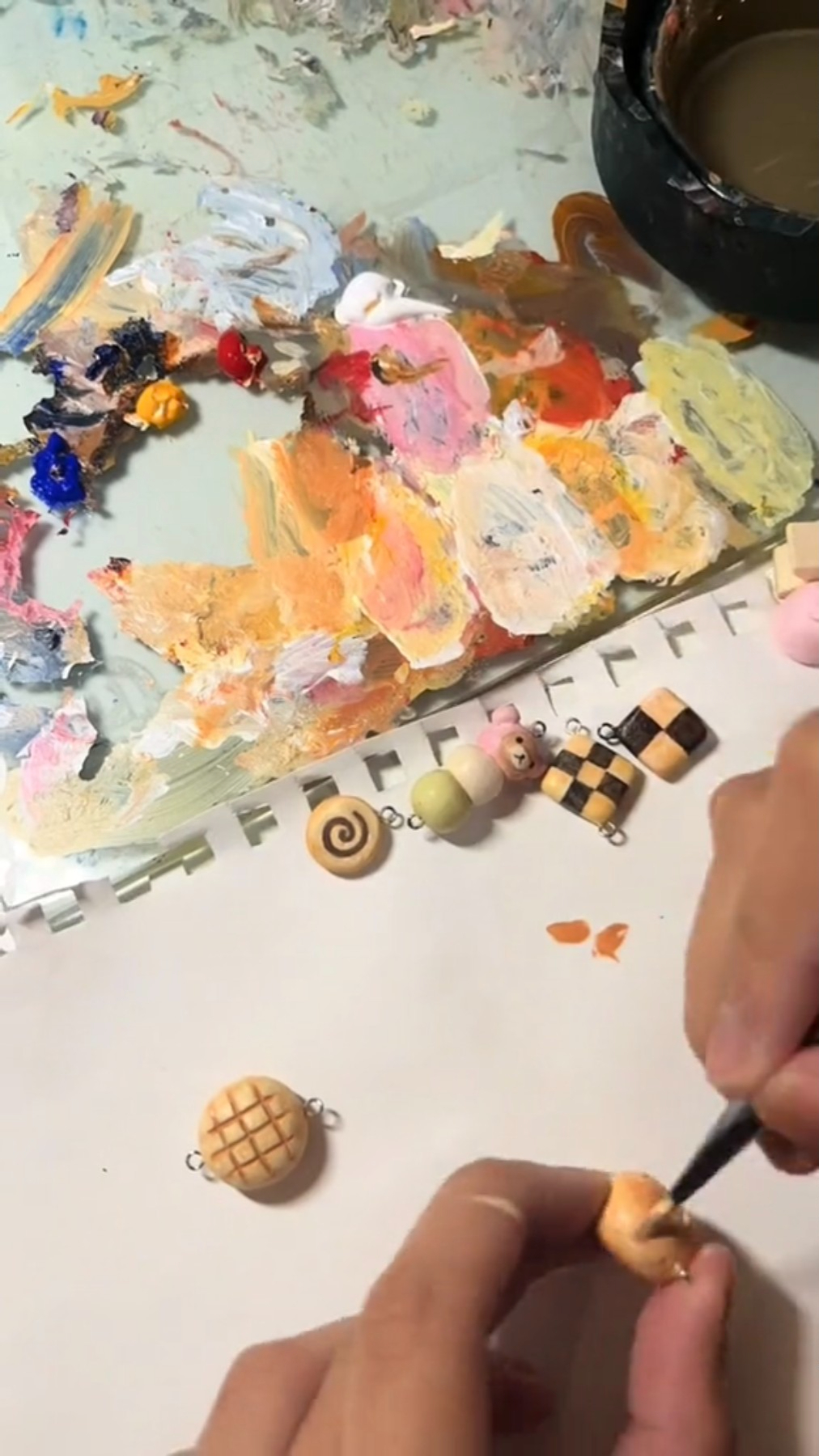 the image shows a person painting miniature food charms on a table covered in art supplies. the charms, possibly made of polymer clay, depict various treats like a croissant, a checkerboard cookie, a swirl lollipop, and more. the artist is using a fine brush to carefully add details. a palette with various paint colors is visible in the background, along with a container and some painted charms waiting to be finished.