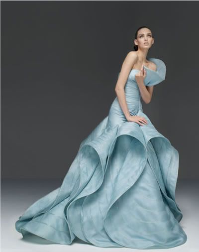 a woman in a stunning light blue gown with ruffles and a cinched waist poses elegantly against a grey background. the gown flows around her in soft fabric folds, creating a  romantic and luxurious feel. the woman's style is sophisticated and gorgeous, embodying both elegance and effortless beauty.