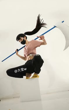 a young woman is posing with a blue pole in her hand. she is wearing a black mask and black clothes. she is standing on a white box. the background is white.