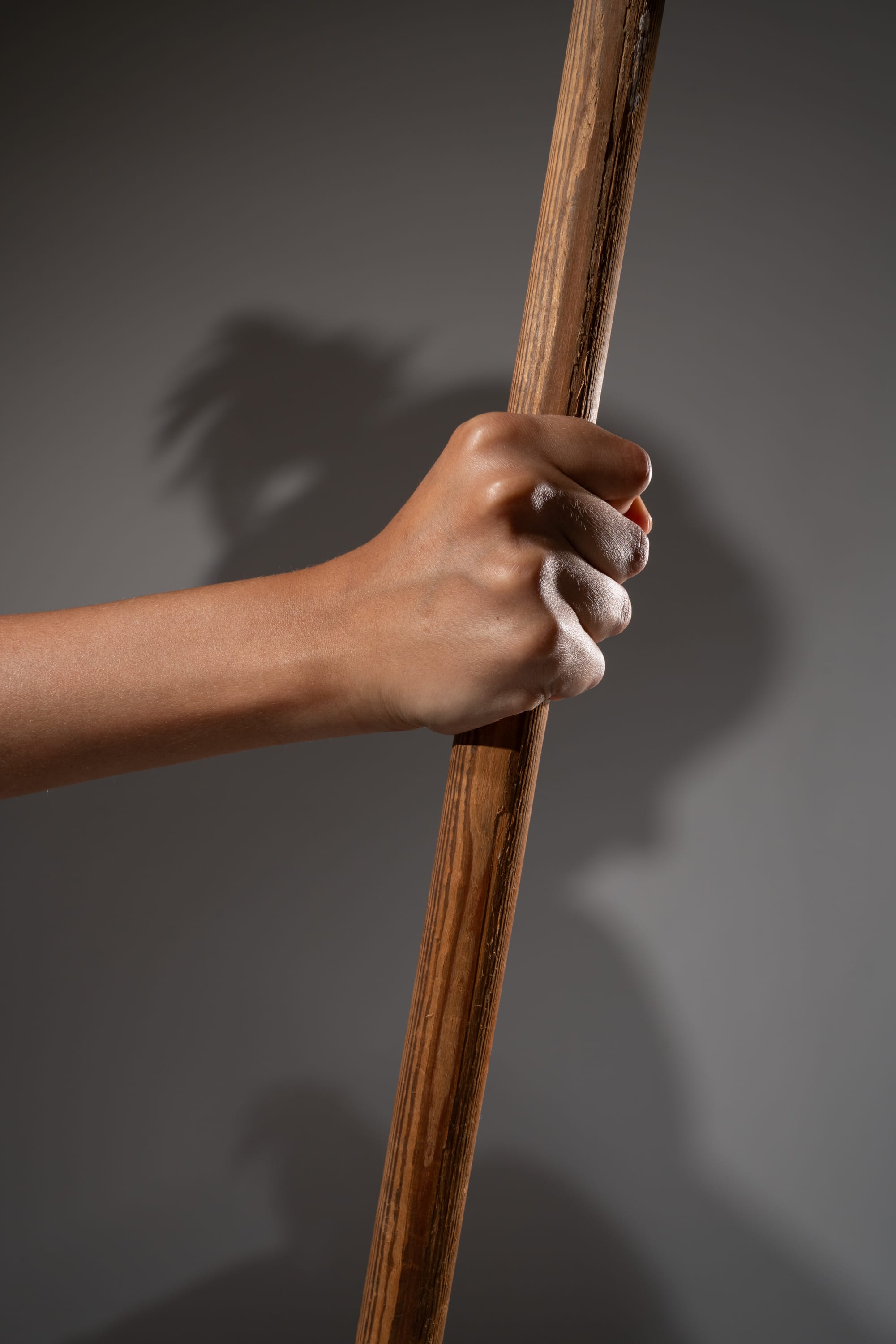 a hand tightly grasps a wooden staff. the hand and arm are illuminated by studio lighting, highlighting the tension and grip. a shadow of the hand and staff falls onto the background, adding to the dramatic effect.