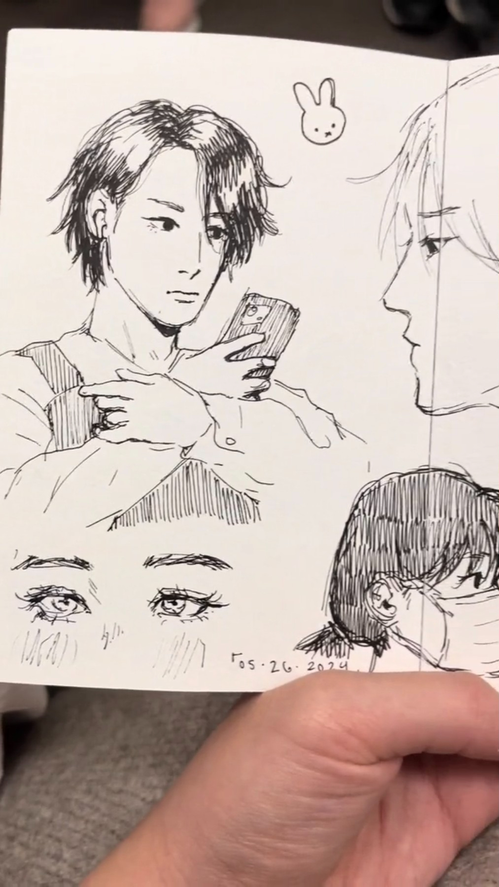 a hand drawn sketch of a man with dark hair looking down at a phone he's holding in his hand. there is a bunny doodle in the top right corner of the page. the top right corner of the page also features the face of another man with short white hair. the page has the date written in the bottom right corner. the bottom left corner has a hand drawn eye. the page is being held in the hand of a person.