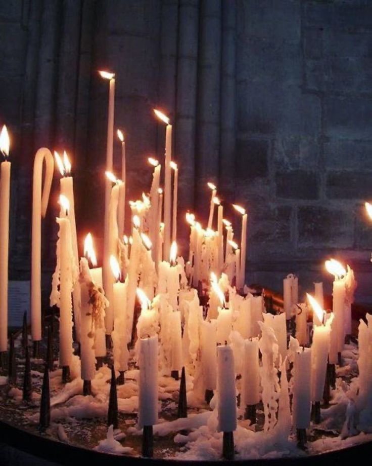 the image shows a dense collection of white candles burning brightly on a metal candle stand with spikes. the candles are of varying heights and have significant wax drippings. the background reveals a dark, stone wall, possibly inside a church or gothic building. the overall atmosphere is dimly lit and evocative of religious rituals and prayer.