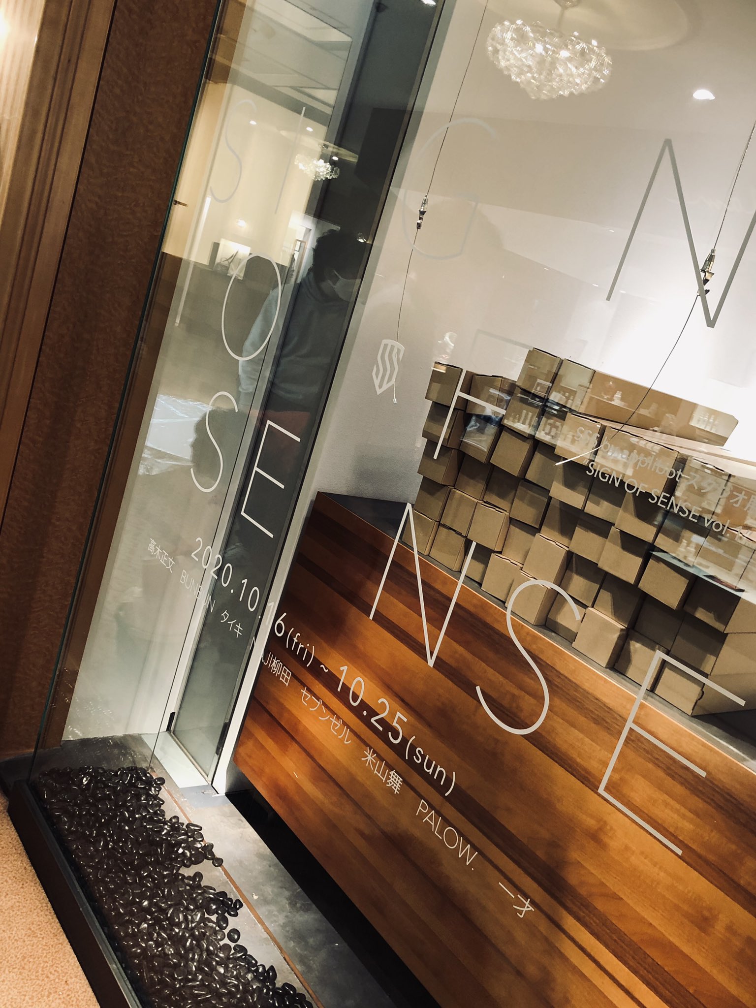 this image shows the exterior of a storefront with large glass windows. the name "sign of sense" is visible on the glass, along with japanese and english text indicating an exhibition or popup event taking place inside.  several stacks of cardboard boxes are visible, perhaps containing merchandise or artwork. at the bottom of the window, there's a decorative element filled with coffee beans. a person's reflection can be seen in the glass. the overall aesthetic is minimalist and modern.
