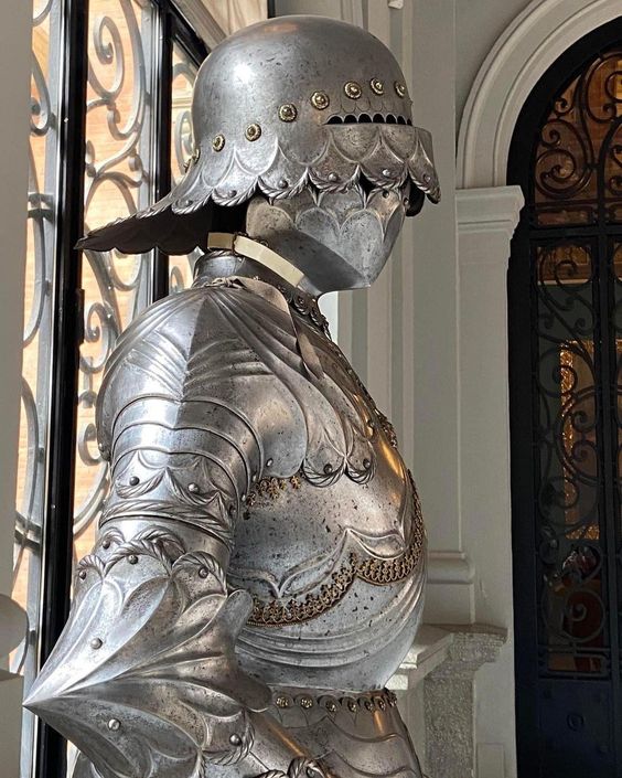 a closeup view of an empty and ornate suit of medieval armor, displayed on a stand. the armor is made of polished metal and features intricate details. the helmet, with its distinctive shape and rivets, is particularly striking. the background suggests an elegant interior setting, possibly a museum or historic building.