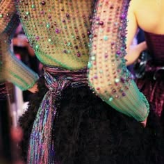 a woman in a sparkly, green, mesh dress with a black skirt and a purple scarf tied around her waist. she is standing in front of a blurred background of other people and the focus is on the dress and its intricate details.