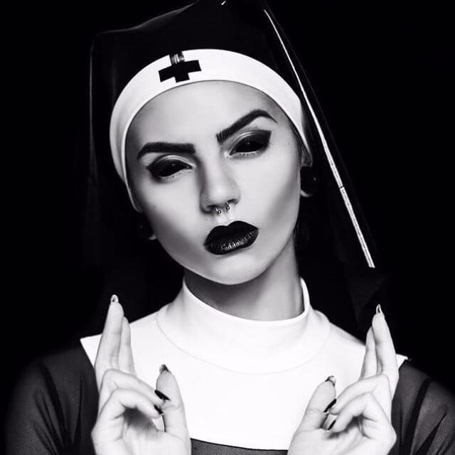 a woman dressed as a nun, with dark makeup, black clothing and a white cross on her head. she is praying, with her hands clasped in front of her. her eyes are shadowed and her lips are painted black. the image has a dark and gothic feel to it.
