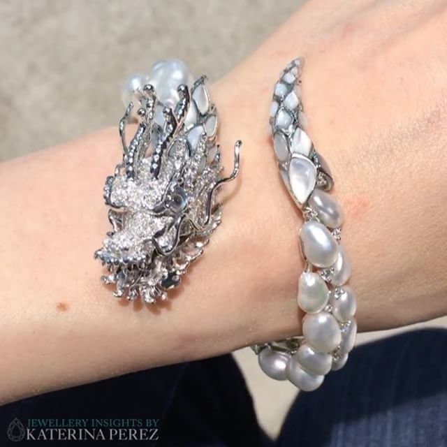 the image shows a closeup of a woman's wrist wearing a striking silver dragonshaped cuff bracelet adorned with pearls and diamonds. the dragon head is detailed with intricate designs and positioned on the top of the wrist. the cuff extends with pearls and silver accents down the arm. the image is credited to "jewellery insights by katerina perez."