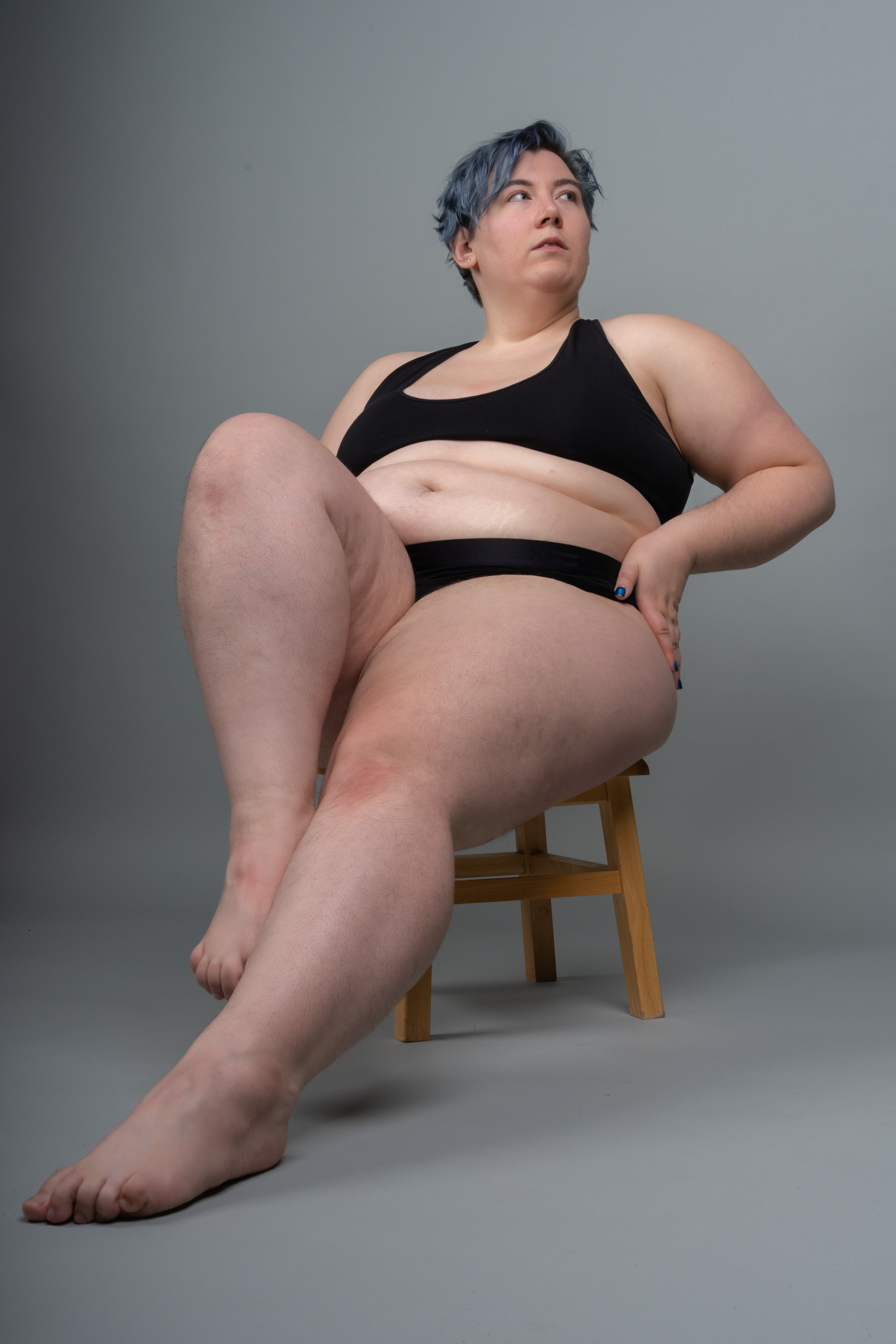a portrait of a plussize woman with blue hair, wearing black underwear, sitting on a wooden stool with her legs crossed. she is looking away from the camera. the background is a solid grey color.
