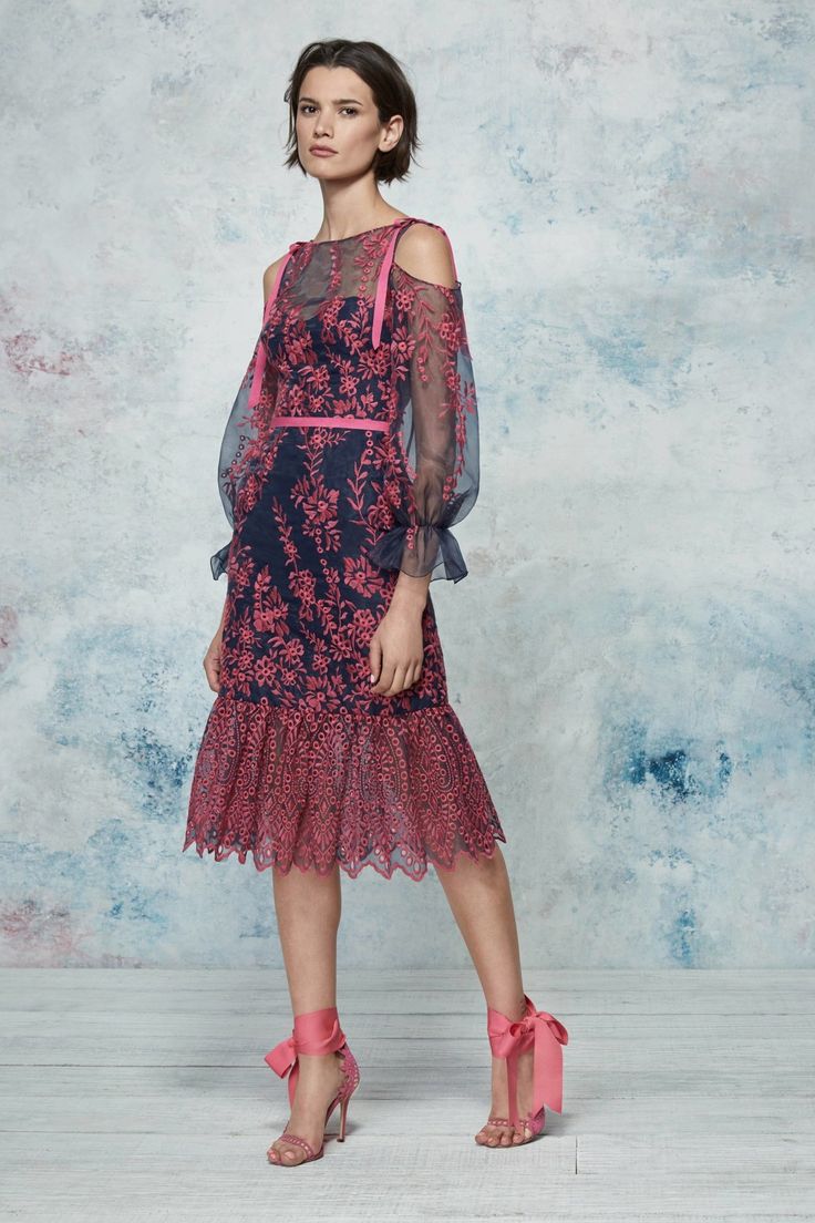 a full shot of a female model showcasing a navy blue and pink embroidered midi dress with cold shoulders and sheer long sleeves. the dress has a ruffled hem and is cinched at the waist with a pink ribbon. the model is wearing pink heels with ribbon ties and has short brown hair. the background is a mottled blue and grey textured wall. the image is likely from a fashion photoshoot or lookbook.