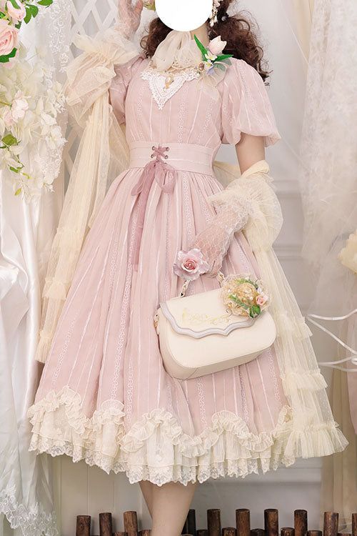 a woman stands indoors modeling a pink, frilly, floor length dress with lace trim. the dress has a sweetheart neckline and puffed sleeves and is cinched at the waist with a ribbon. the woman holds a small, white purse.