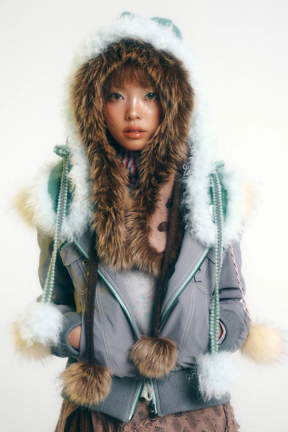 a young woman, possibly a model, is captured in a studio shot against a plain white background. she's wearing a stylish, light grey winter jacket with a large, fluffy hood and accents of light mint green. the jacket features generous amounts of faux fur trim, including a large furlined hood, a fur scarf, and several small pompoms attached to drawstrings and the cuffs. the overall aesthetic is decidedly cute and "kawaii," blending pastel shades with playful textures. the girl's expression is calm and direct, drawing attention to her soft features and the detail of the coat.  she is wearing a delicate lacelike garment underneath the jacket.