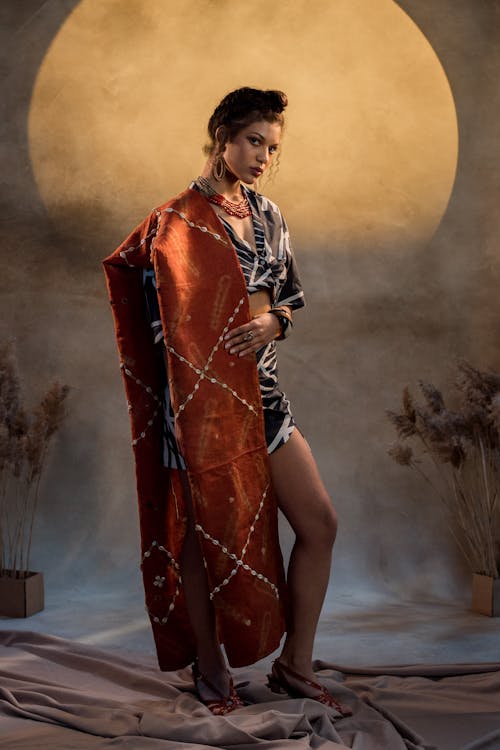 a fulllength shot of a fashionable woman posing in a studio. she's wearing a striking rustcolored shawl or wrap adorned with intricate details, paired with a black and white printed short dress. her accessories include a necklace, earrings, and stylish sandals. the background is a textured wall with a large, softly lit circular area behind her, enhancing the dramatic effect.  she stands confidently with a serious gaze directed slightly offcamera. the overall style evokes a sense of elegance, with elements that hint at cultural or ethnic influences.