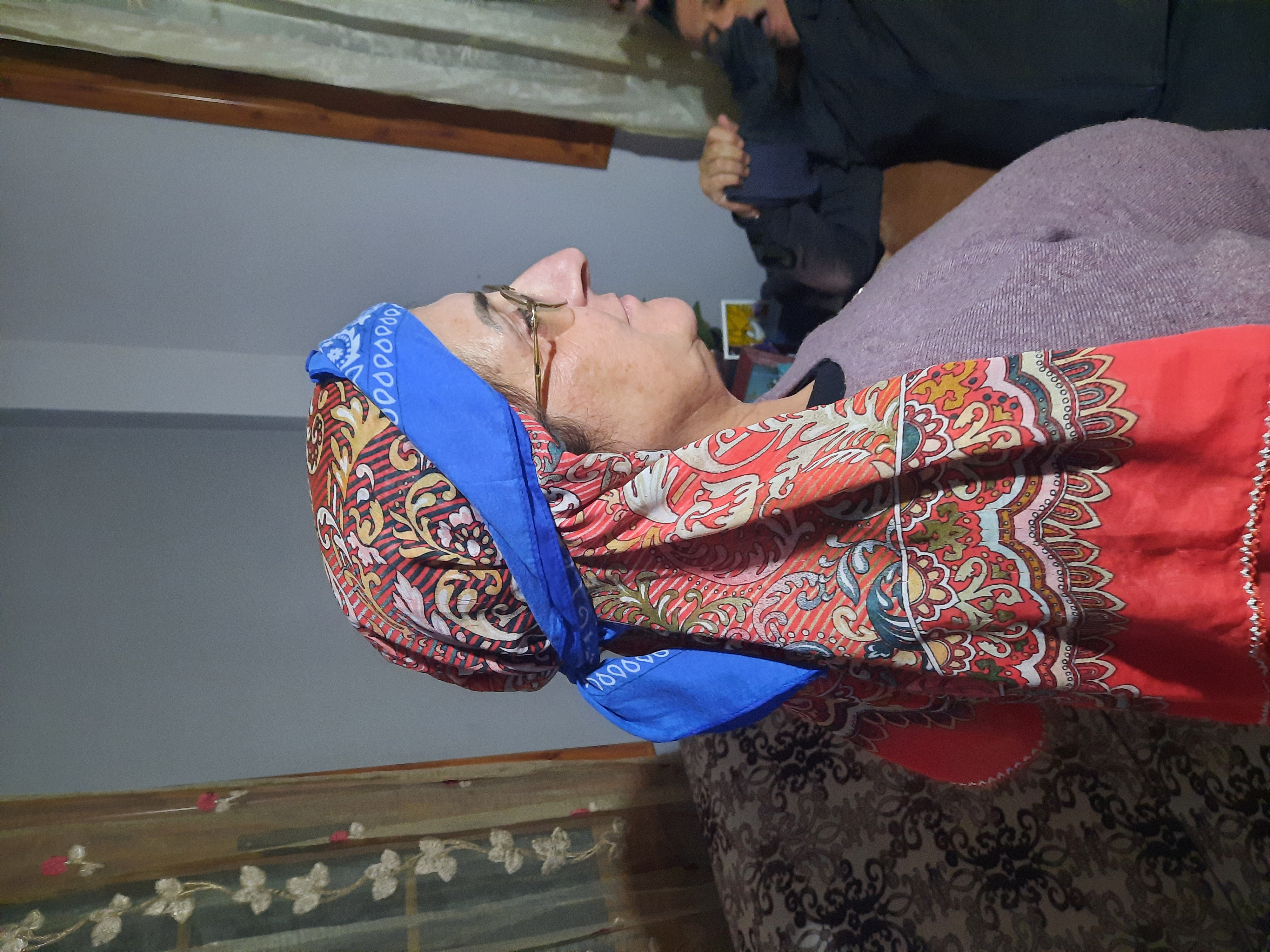 an elderly woman wearing glasses and a patterned headscarf with a blue bandana is seen in profile indoors. she is wearing a light purple sweater and is sitting on a sofa. another person is partially visible in the background.