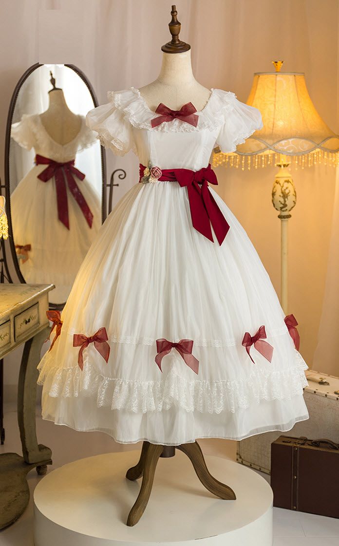 a white vintagestyle dress is displayed on a mannequin. the dress features red bows and ribbons, lace trim, and a ruffled skirt. the dress is shown in a welllit room with a mirror and a lamp in the background.