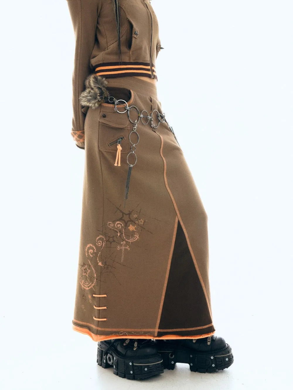 closeup view of a woman's outfit featuring a long, brown corduroy skirt with an asymmetrical design and orange stitching details. the skirt has a dark brown panel down the side and an embellished design. she is also wearing a matching brown longsleeved jacket with orange stripes at the waist, a metal chain belt, and chunky black platform boots. the style evokes a gothic or alternative fashion aesthetic. the image is taken against a white background, which helps to highlight the details of the outfit.