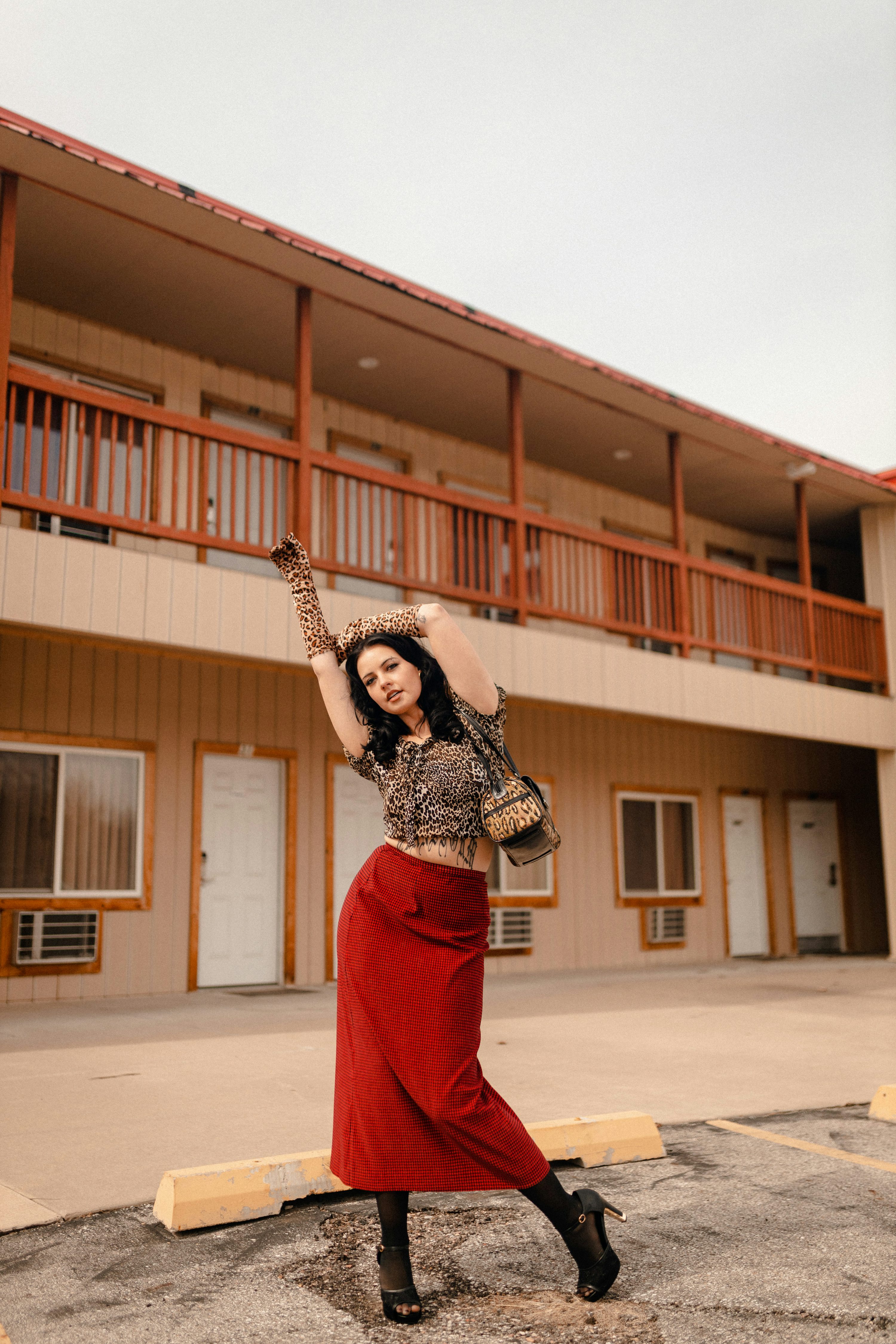 a stylish woman poses confidently in a motel parking lot. she is wearing a red maxi skirt, a leopard print top, and long gloves. she carries a handbag and is wearing high heels. the motel building is a light beige color with wooden balconies. the overall aesthetic is retro and vintage inspired.