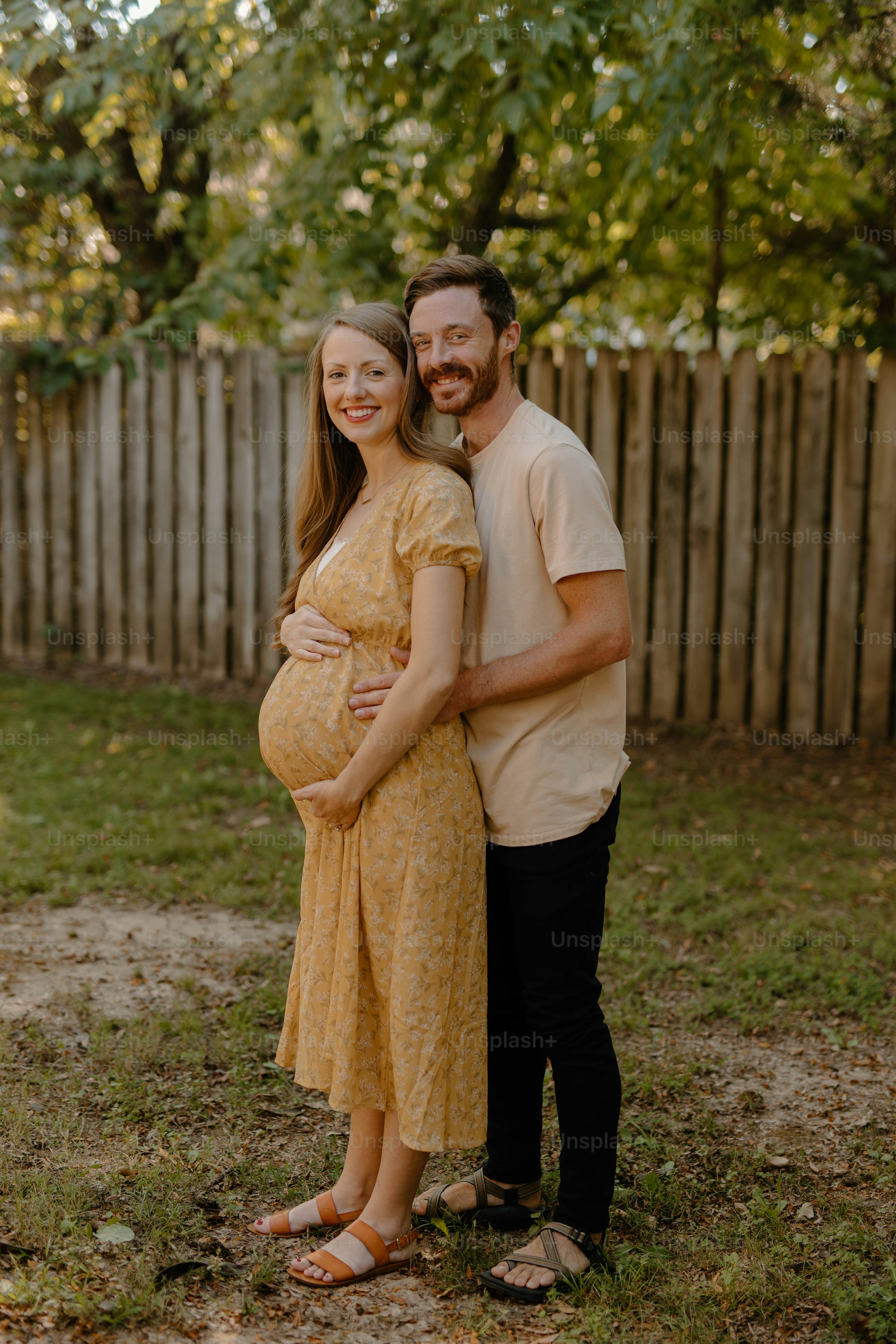 a pregnant woman in a yellow dress stands close to her partner, both smiling happily. they are outdoors in a garden setting next to a wooden fence. the sun is shining, and the overall mood is joyful and loving.