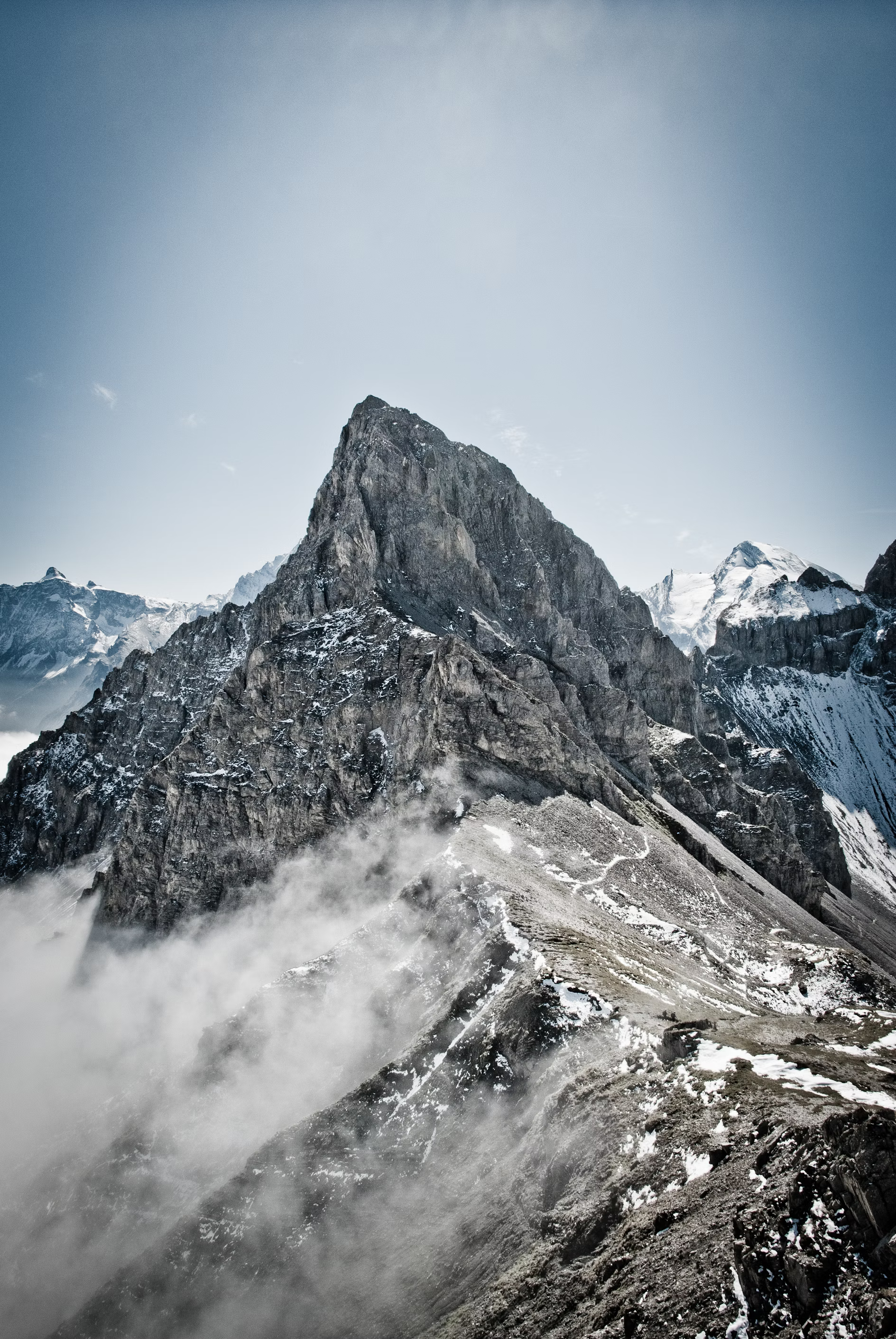 the image shows a majestic mountain peak partially shrouded in fog. the rocky slopes are dusted with snow, and a winding path suggests a challenging climb. the sky is clear and blue, adding to the sense of grandeur and isolation.