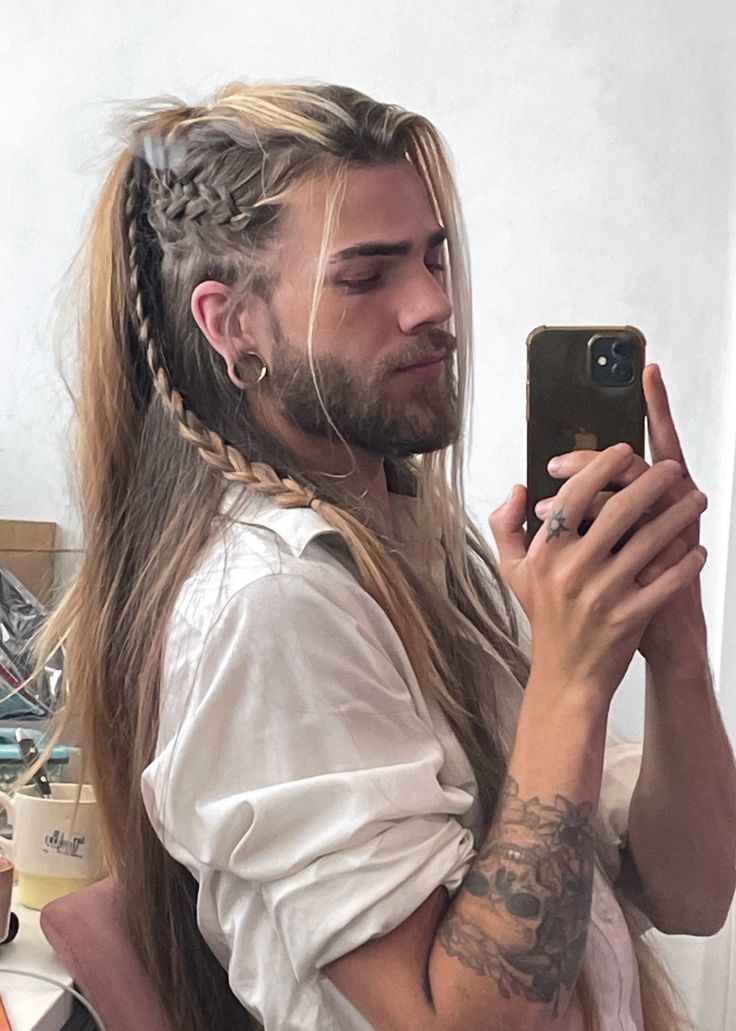 a young man with long blonde hair that is braided is taking a selfie with his phone. he has a beard and a tattoo on his wrist. he is wearing a white shirt.