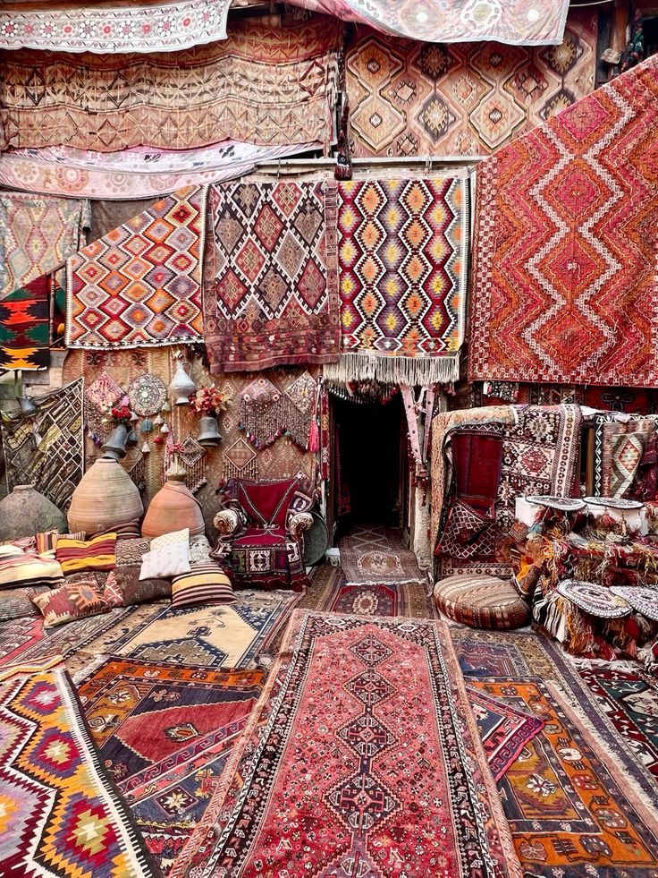 the image shows a shop or bazaar filled with a vast collection of carpets, rugs, and textiles. the walls are covered in hanging carpets with intricate geometric patterns and vibrant colors like red, brown, orange, and yellow. the floor is also layered with carpets and rugs of various sizes and designs. cushions and pillows are scattered around, and there's an antique chair in the center. the scene evokes a sense of richness, history, and traditional craftsmanship.