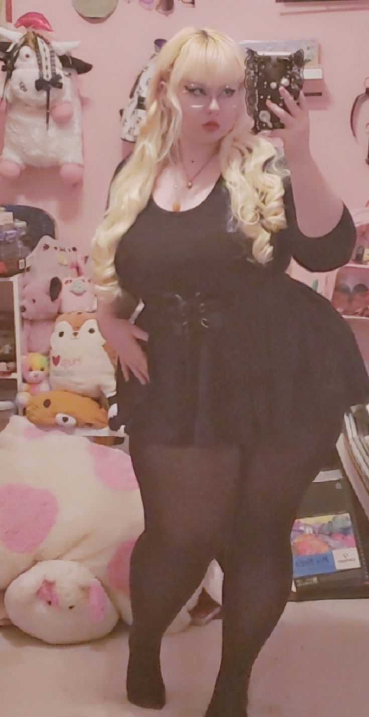 a blonde woman with black framed glasses poses for a selfie in her bedroom.  she is wearing a black dress with a corset, black tights, and has her long blonde hair down.  the woman is standing in front of a white shelf with pink toy dolls and a mirror.  the wall behind her is pink.