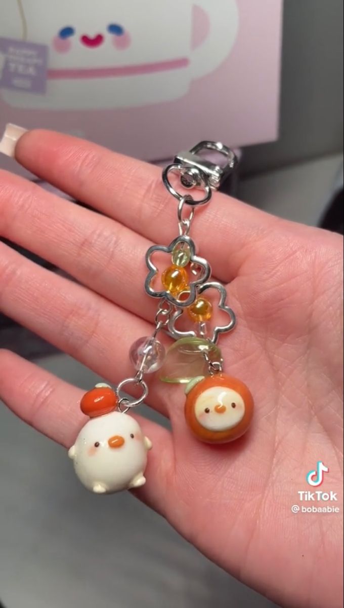 a hand holds a keychain with various charms and beads. the keychain has a silver clasp and flowershaped charms with orange beads, clear beads, and two chick charms. one chick is white with an orange hat, and the other is orange. the background is a light pink tea cup.