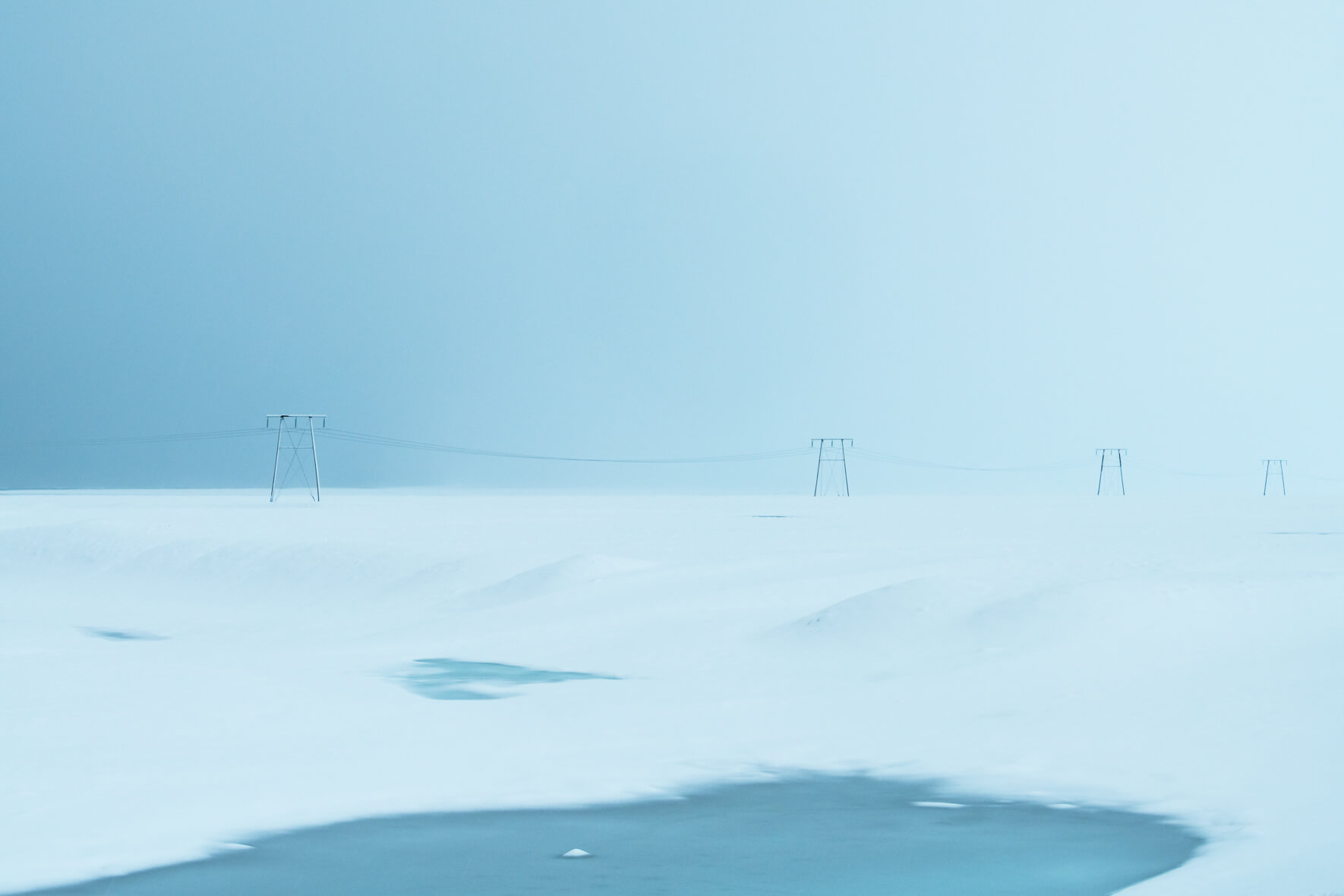 a panoramic landscape photograph featuring a vast expanse of snowcovered terrain. the horizon is visible in the distance, with a faint blue sky above. four tall power lines stand in a row, silhouetted against the snowy landscape. their metal structures and wires are stark against the white background, emphasizing the manmade presence in this otherwise pristine wilderness. the scene is characterized by a sense of stillness and solitude, highlighting the vast and isolated nature of this remote location.