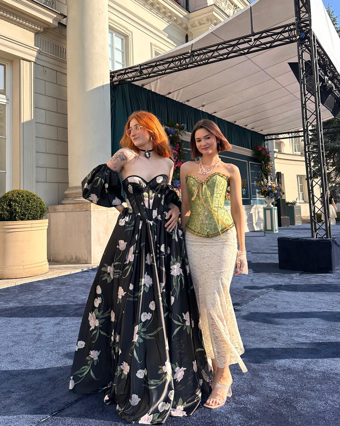 two young women stand on a pathway outside a grand palace.  one woman wears a long, black floral gown with a fitted bodice, and the other wears a lime green corset top and a long, ivory lace skirt. they are smiling, and both appear elegant and happy, possibly at a formal event or celebration.  the background features the classic architecture of the palace and a temporary event structure. the overall atmosphere is stylish and sophisticated.