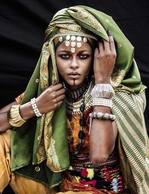 a woman of african descent is shown wearing a traditional green head covering and elaborate jewelry. she has dark skin, dark hair, and wears a colorful outfit with gold jewelry. she is posing with one hand near her face and the other hand resting on her lap. the woman is looking at the camera and has a serious expression on her face. the photo is taken in a dimly lit studio setting. the background is blurred and black.