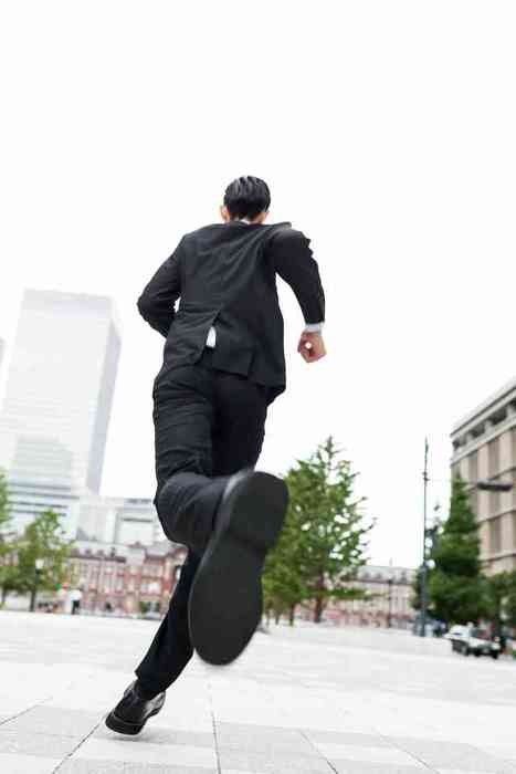 a man in a black suit is running away from the camera. the photo is taken from a low angle, giving the impression of speed and urgency. the man is in focus, while the background of buildings and trees is slightly blurred. the overall palette is muted, with grays and blacks dominating.