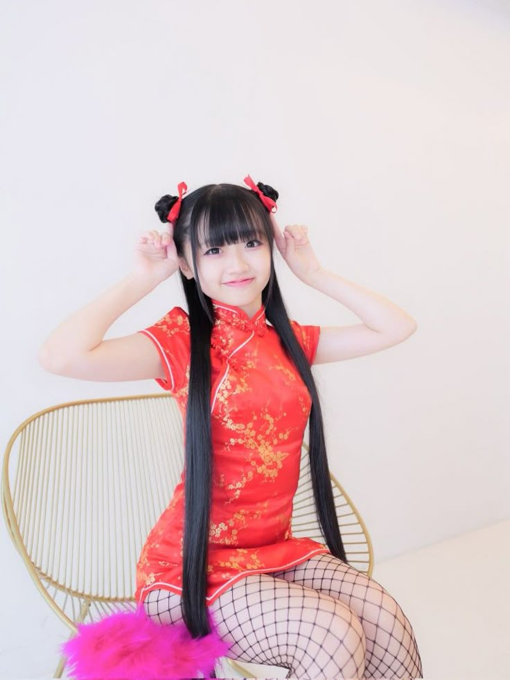 a young asian woman with long black hair and bangs, styled in pigtails with red bows, is smiling and sitting in a gold chair. she is wearing a short, red qipao or cheongsam dress with gold floral designs and fishnet stockings. her hands are raised near her head in a playful manner. the background is a plain white wall. a pink, fluffy item is visible on the seat of the chair.