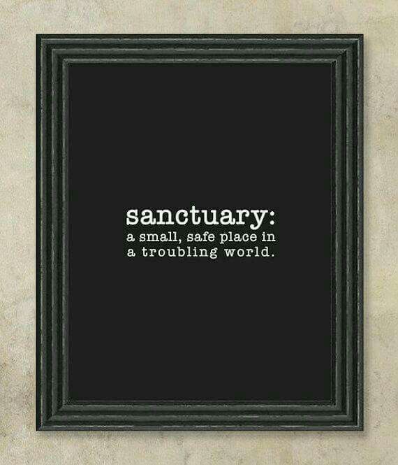 the image shows a framed print against a lightcolored textured wall. the print has a black background with the word "sanctuary:" in white, followed by the definition "a small, safe place in a troubling world." the frame is a dark, ornate style. the overall effect is a decorative piece with an inspirational message about finding safety and peace.