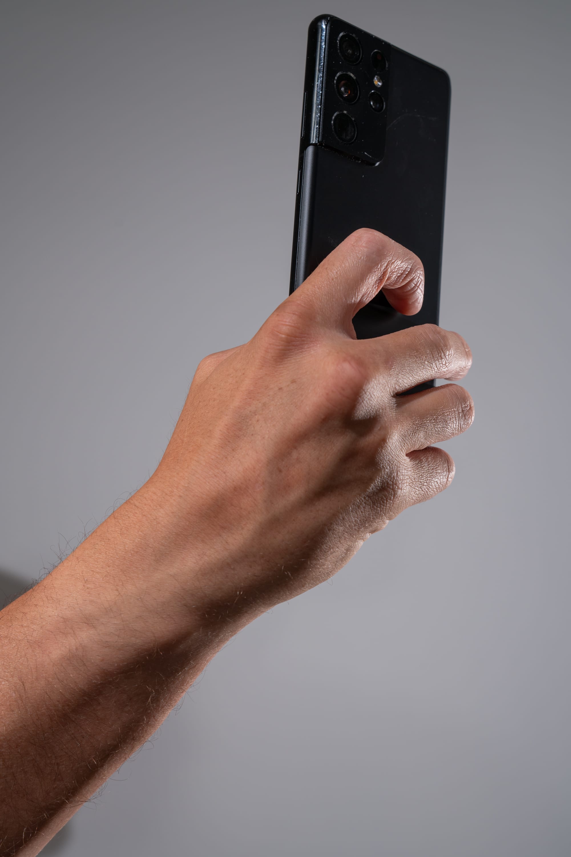 a closeup, lowangle view of a person's hand holding a black samsung galaxy smartphone against a neutral gray background. the hand is positioned so that the index finger is resting on the phone's screen.  the smartphone's prominent multilens camera array is visible on the back of the device.