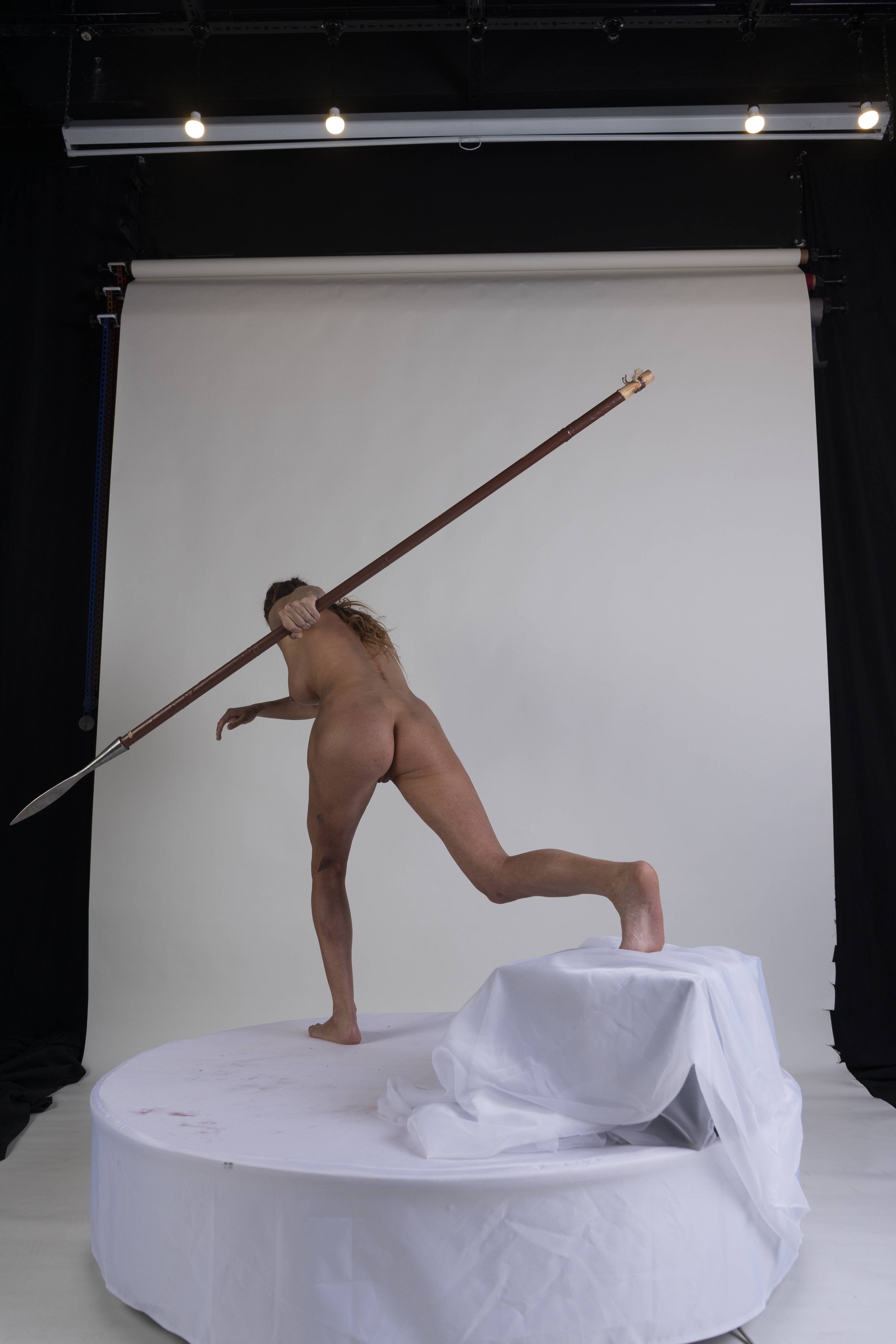 a nude woman stands on a white platform with draped fabric, holding a spear. the scene is set in a studio with a white backdrop and black curtains on either side. lighting fixtures are visible at the top of the frame. the woman is in motion, appearing to be throwing or wielding the spear. her back is facing the viewer.
