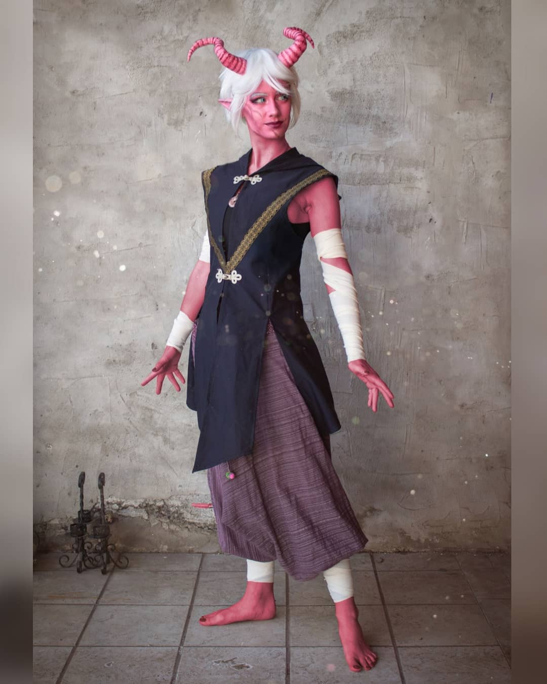 a fullbody shot of a female cosplayer portraying a tiefling character, likely from dungeons & dragons.  she has white hair, pink skin, and pink horns. her costume consists of a dark vest over a purple skirt,  wrist wraps, and barefoot.  the background is a textured grey wall. the overall style is fantasythemed, and the image is welllit, appearing to be a professional studio shot.