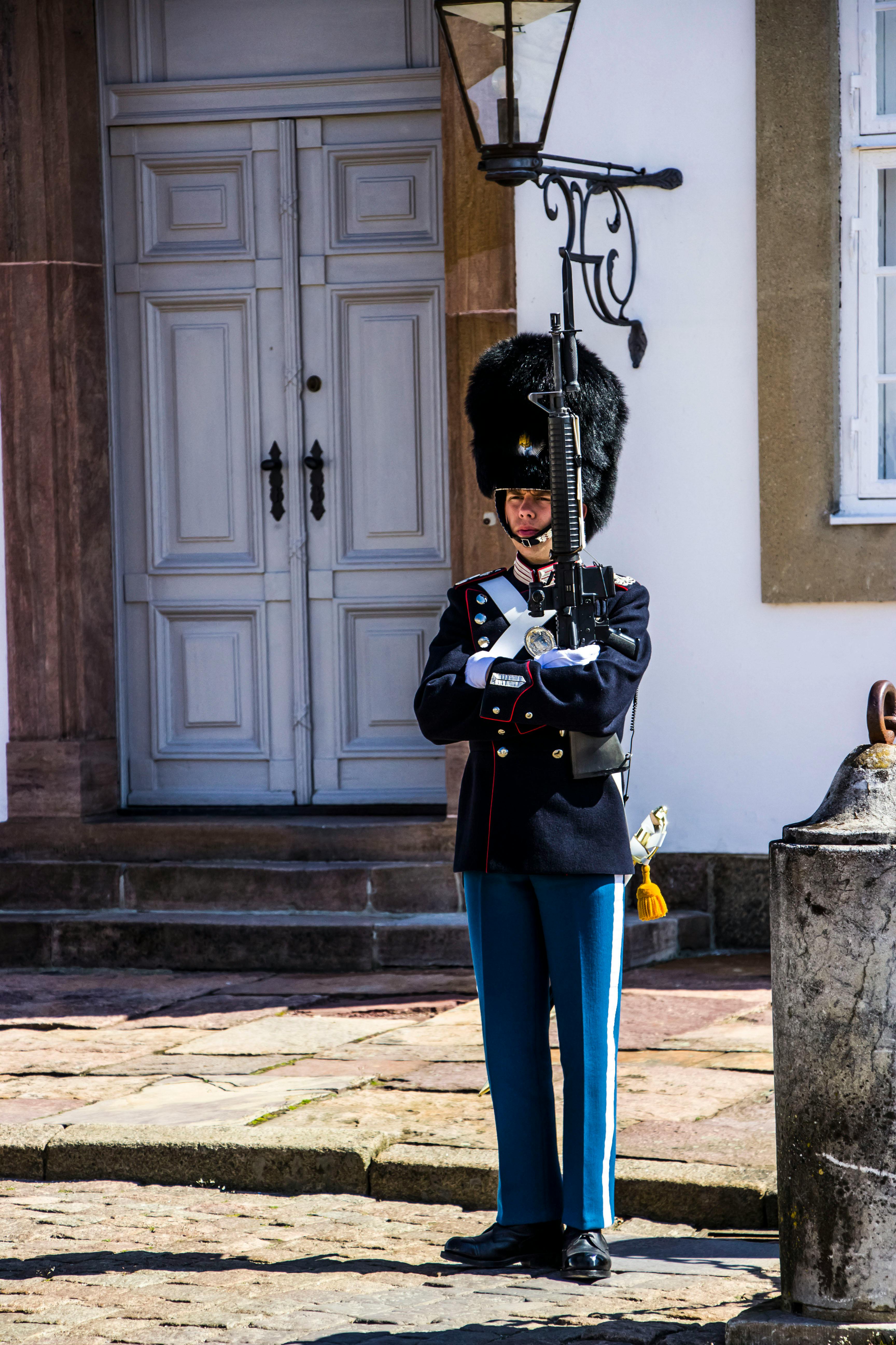 a danish royal guard soldier stands at attention in front of a white door, holding a rifle. the soldier is wearing a traditional dark blue uniform with a bearskin hat. the setting appears to be a historical building or castle. the soldier is positioned as a sentry, maintaining security. the image conveys a sense of history, tradition, and military formality.