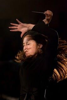 a woman in a black longsleeved shirt stands on a stage with a baton raised in one hand, she is directing an orchestra, her eyes are focused and her face has a serious expression. the stage is dark and lit by dramatic spotlights.  the woman has long brown hair and is wearing a black longsleeved shirt.  she is conducting an orchestra and is focused on her task.