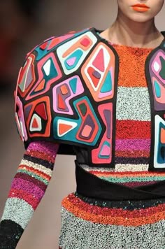 a woman is wearing a stylish and colorful jacket with a geometric pattern. the jacket is made up of vibrant shades of pink, blue, red, and yellow, and it is paired with a striped skirt that adds a touch of classic style. the outfit is completed with a black belt and a pair of shiny sequins that reflect the light in a beautiful way.