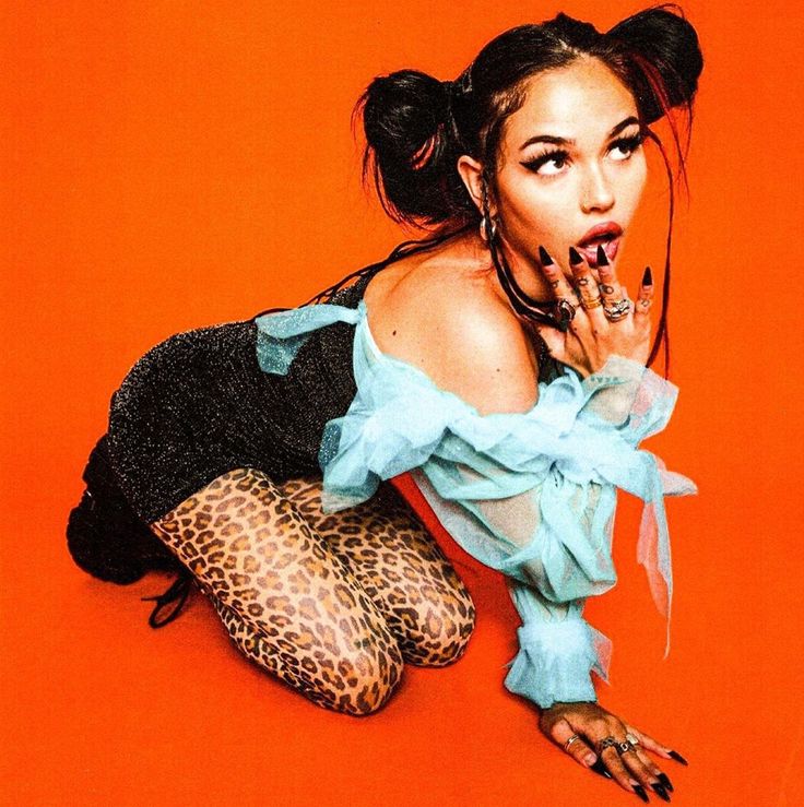 a young woman with dark hair styled in two buns, wearing a black and silver outfit with leopard print tights and a blue sheer top, is sitting on an orange background and looking at the camera. she is wearing a variety of jewelry and has long nails. her pose is sensual and her expression is playful and confident. the lighting is dramatic and the image has a glamorous feel.