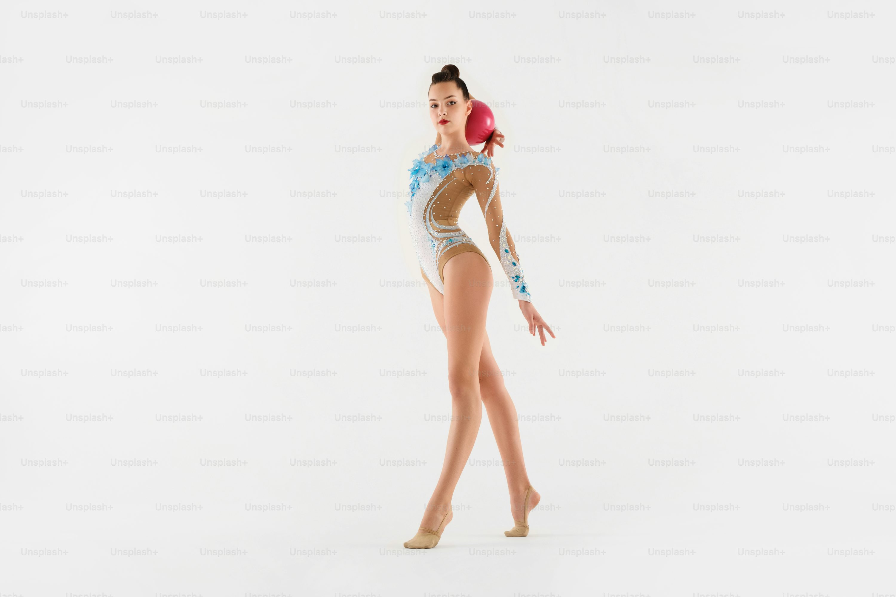 a young female rhythmic gymnast, poised and graceful, performs a pose with a pink ball in her hand against a clean white background. she is wearing a beautiful white and blue leotard and her posture demonstrates both strength and flexibility.  the image showcases her athletic skill and artistry.